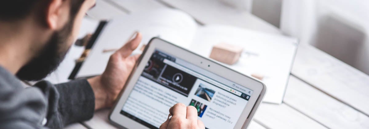 man reading web page on tablet with coffee and open book nearby