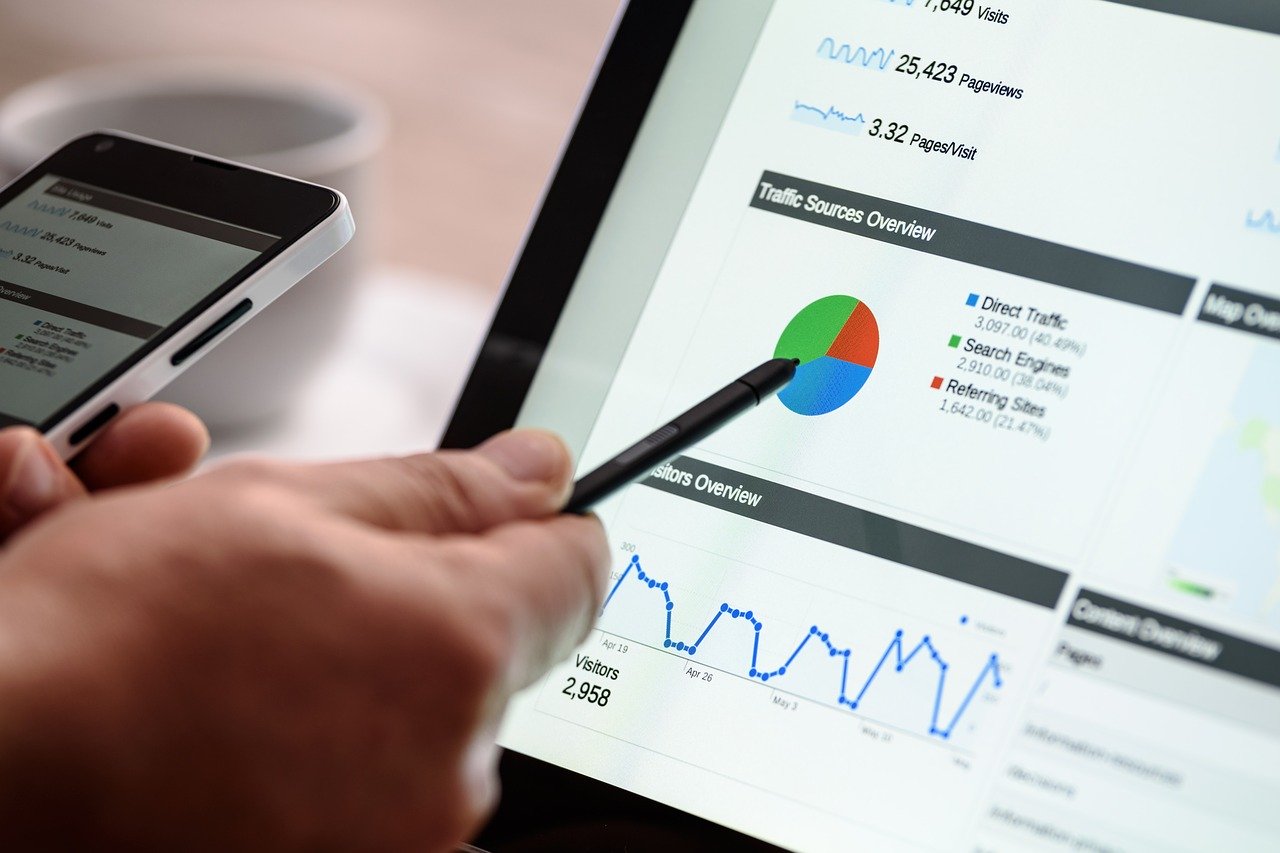 person pointing to pie chart on website analytics overview page on computer monitor, comparing it to data on smartphone 